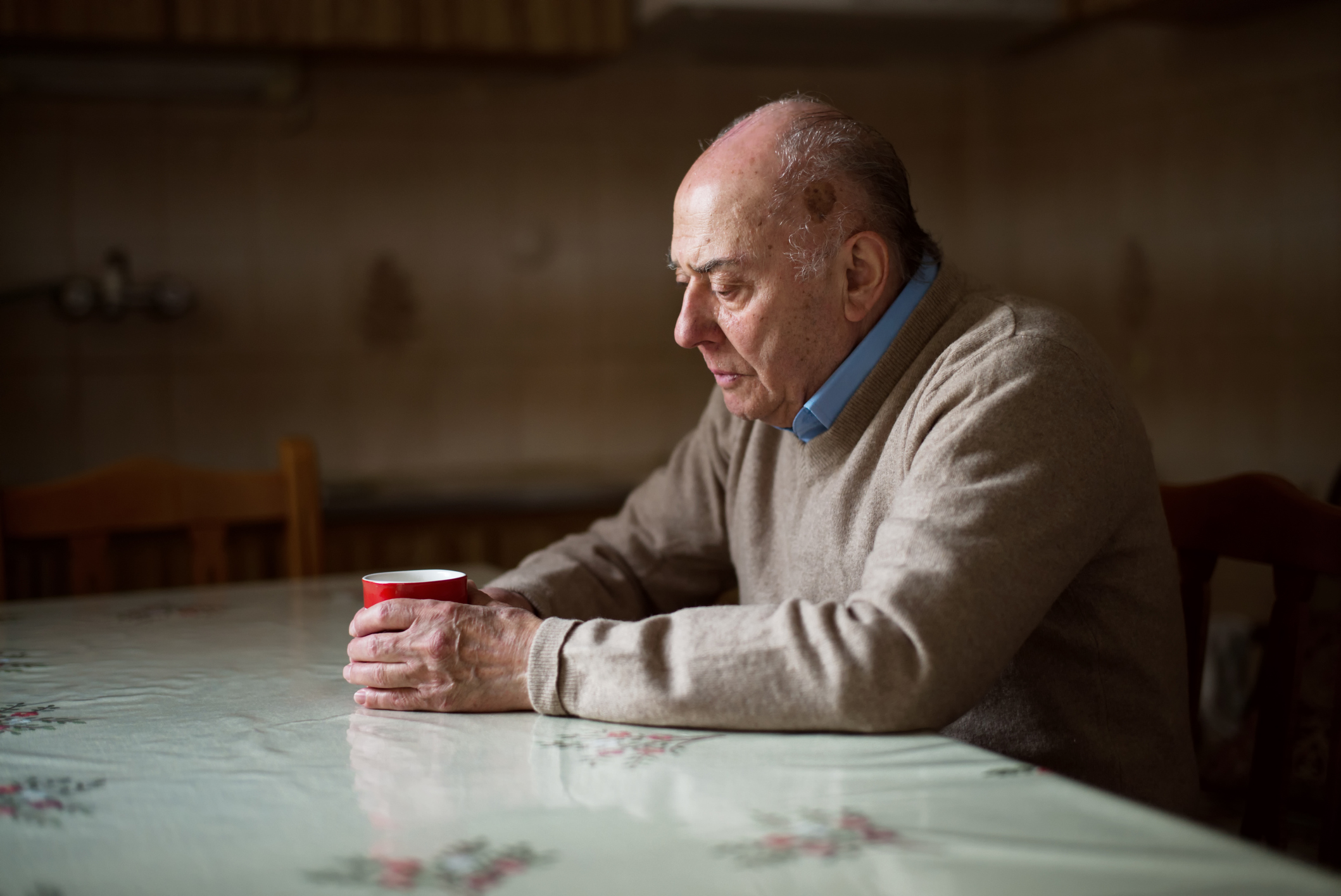 Das Foto zeigt einen älteren Mann, der gebeugt an einem Tisch sitzt und eine Tasse umklammert.