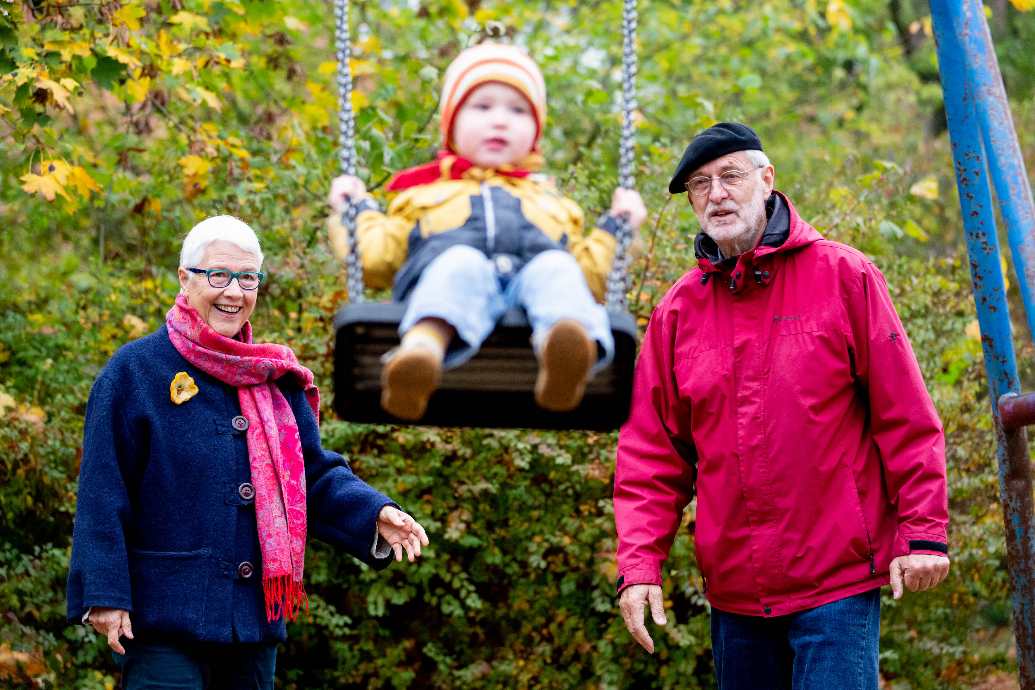 Das Foto zeigt ein Kleinkind auf einer Schaukel, rechts und links davon stehen ein älterer Mann und eine ältere Frau, bei denen es sich vermutlich um die Großeltern handelt